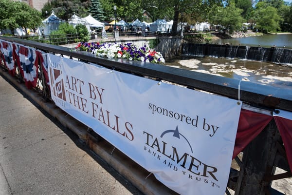 A large banner on the bridge overlooking the river reads, Art byt the Falls sponsored by Talmer Bank and Trust