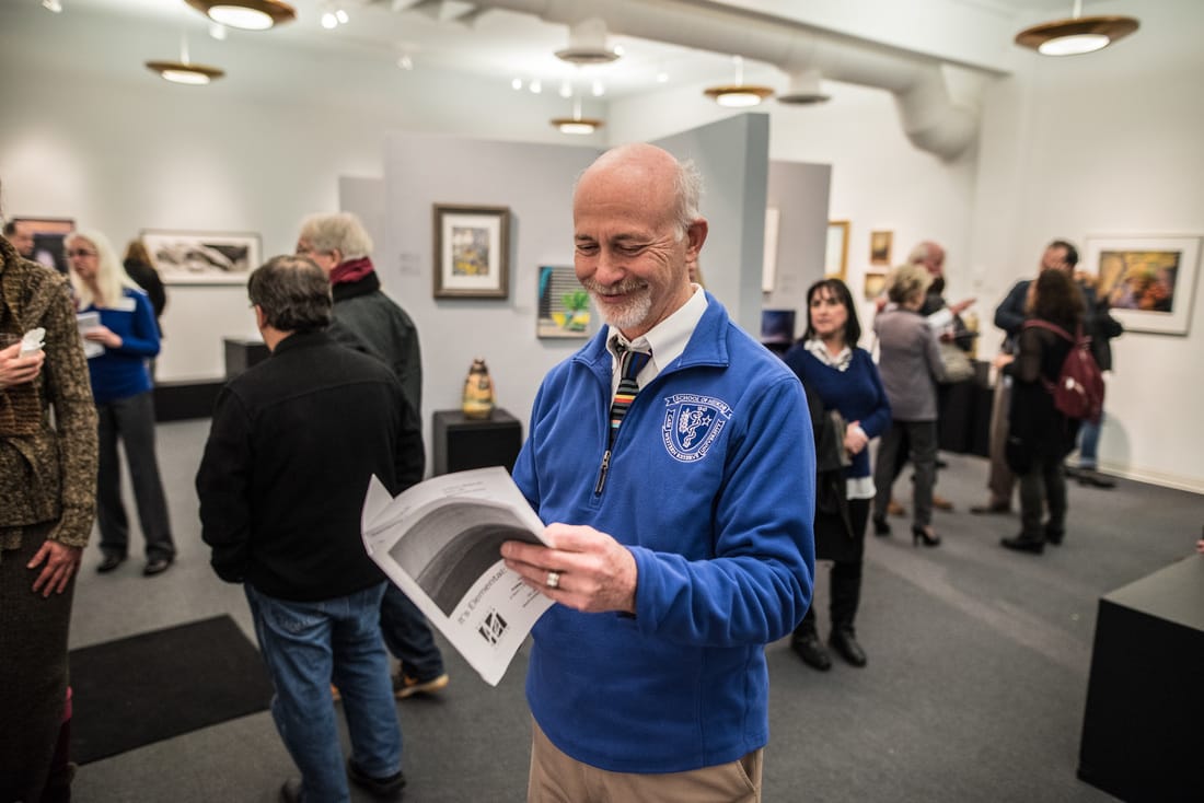 Smiling man reads an exhibit program among groups of visitors in the gallery